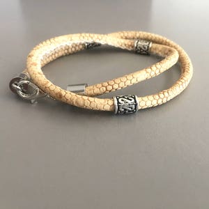 May include: A double-wrap bracelet made of light brown leather with a textured pattern. The bracelet has two silver metal beads with decorative designs.