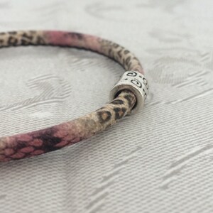 Könnte beinhalten: Ein rosafarbenes und braunes Lederarmband mit einem silbernen Perlenanhänger. Die Perle hat ein dekoratives Design.