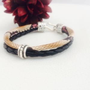 Op de afbeelding: Een dubbele leren armband met een slangenhuidpatroon en een zilveren sluiting. De armband is gemaakt van twee leren strengen, een zwarte en een bruine met een slangenhuidpatroon.