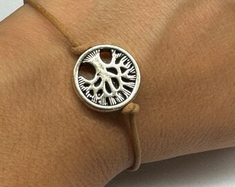 May include: A silver-colored tree of life charm bracelet. The charm is circular with a detailed tree design. The bracelet has a thin, light brown cord that adjusts to fit the wrist. The bracelet is a simple, elegant piece of jewelry.