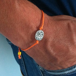 May include: A close-up of a wrist wearing an orange string bracelet with a silver charm featuring a floral design.