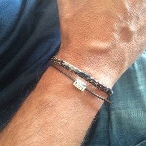 Könnte beinhalten: Zwei Armbänder an einem Handgelenk. Das obere Armband ist ein geflochtenes Lederarmband mit einem silbernen Anhänger. Das untere Armband ist eine braune Lederschnur.