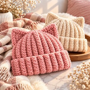 May include: Two crocheted beanies featuring cat ears, one in a soft pink and the other in a cream colour. Both hats are crafted with a thick, ribbed knit texture and include a wide, folded brim. The beanies are positioned together, showcasing the distinct pointed ear details and the consistent yarn construction of each piece.