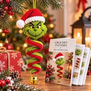 Puede incluir: Un adorno de ganchillo con la cabeza de un personaje verde con un gorro de Papá Noel rojo y blanco, unido a un cuerpo en espiral rojo y verde que termina en un cascabel dorado. Junto a él, un folleto dice CROCHET PATTERN PDF DOWNLOAD.