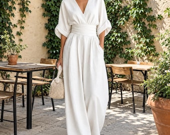 May include: A white jumpsuit featuring a deep V-neckline, wide elbow-length sleeves with folded cuffs, and a wide gathered waistband. The garment has long, wide-leg trousers with side pockets and is paired with a small, textured beige handbag.