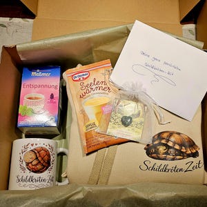 Könnte beinhalten: Eine Geschenkbox mit einer Packung Meßmer Entspannung Tee, Dr. Oetker Seelenwärmer Vanille-Pudding, einer weißen Tasse mit Schildkröten-Illustration und dem Text Schildkröten-Zeit, einem kleinen Beutel mit Herzanhänger sowie einer Notiz.