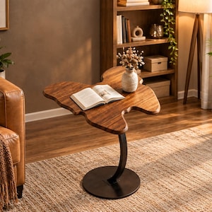 May include: A side table with a natural wood grain top shaped like a butterfly, supported by a curved black metal stem and a circular black metal base. An open book rests on the tabletop, and a small ceramic vase with dried flowers sits nearby.