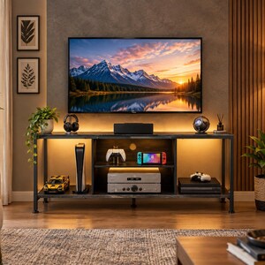 May include: A rectangular media console with a dark metal frame and wooden shelves, featuring integrated warm LED lighting. The unit holds a gaming console, a handheld gaming device, controllers, and audio equipment, positioned beneath a wall-mounted television.