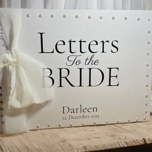 Könnte beinhalten: Ein weißes, spiralgebundenes Album mit dem in Schwarz gedruckten Text Letters To the BRIDE, Darleen und 22. Dezember 2025. Der Einband ist mit einer weißen Stoffschleife auf der linken Seite und kleinen weißen Perlen an den Rändern verziert.