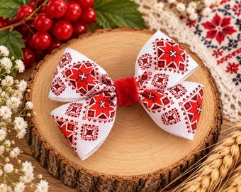 May include: A white hair bow made of ribbed fabric featuring a repeating geometric pattern in red and black. The center of the bow is cinched with a solid red velvet band. The design includes intricate star-like shapes and small cross motifs.
