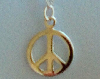 May include: A gold-colored peace sign pendant necklace. The pendant is a classic peace symbol design, suspended from a delicate silver chain. The necklace is displayed against a light blue background.