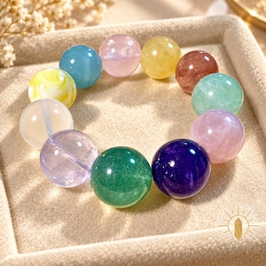May include: A circular bracelet composed of eleven smooth, spherical beads in various colors including blue, pink, yellow, brown, green, purple, and translucent white, arranged in a continuous loop on a soft, textured surface.