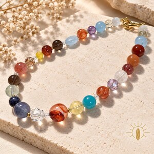 May include: A beaded bracelet featuring a variety of round, polished gemstones in shades of blue, red, orange, yellow, purple, and clear crystal. The beads are strung together with gold-toned metal accents and a gold-toned lobster claw clasp.