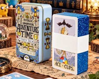 Op de afbeelding: Een lichtblauw metalen blik met de tekst A TOTALLY UNOFFICIAL ADVENTURE TIME TAROT DECK BY KATHERINE HILLIER en een set tarotkaarten met blauwe glitterranden, bijeengehouden door een witte papieren band, met de Aas van Kelken kaart.