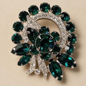 Op de afbeelding: Een ronde broche met een grote, gefacetteerde donkergroene kristal in het midden, omringd door kleinere ronde donkergroene kristallen en marquise-geslepen groene stenen. Het ontwerp bevat zilverkleurige metalen accenten met heldere kristallen.