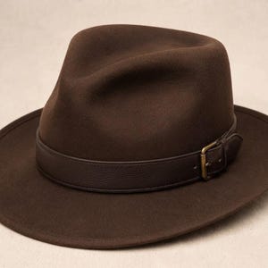 May include: A dark brown felt fedora hat featuring a pinched crown and a wide brim. The hat is accented with a dark brown leather band that wraps around the base, secured by a small, rectangular, antique brass-toned metal buckle on the side.