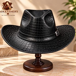 Op de afbeelding: Een zwarte fedora hoed met een textuur van krokodillenleer, voorzien van een bijpassende zwarte band met een zilverkleurige metalen gesp. De hoed staat op een donkerbruine houten standaard. Het logo ADLEATHERS THE TOUCH OF LEATHER is zichtbaar.