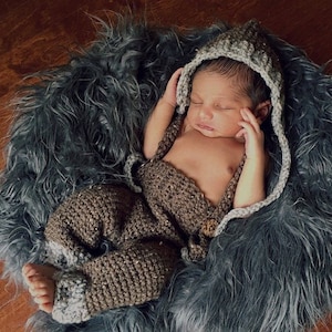 Peut inclure: Un nouveau-né endormi, vêtu d'un ensemble marron en crochet avec une capuche assortie et des bretelles. Le bébé est allongé sur un tapis en fausse fourrure grise.