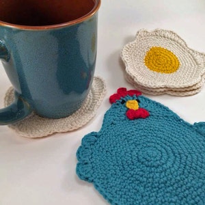 Puede incluir: Tres posavasos de ganchillo, uno con forma de huevo frito, otro con forma de pollo y otro redondo liso. El posavasos con forma de pollo es azul turquesa con una cresta roja y un pico amarillo. El posavasos con forma de huevo es blanco con una yema amarilla. El posavasos redondo es blanco.