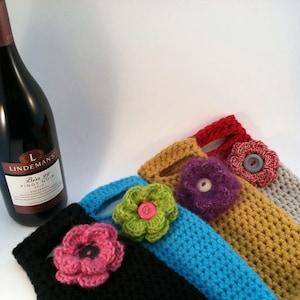 May include: Five crocheted wine bottle holders in different colors, each with a crocheted flower embellishment. The holders are in black, turquoise, mustard yellow, gray, and red. The flowers are in pink, green, purple, and white.