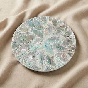 May include: A circular decorative plate featuring a mosaic surface composed of iridescent abalone shell fragments. The shell pieces display a spectrum of shimmering teal, green, and pearlescent white tones arranged in a radial pattern across the disc.