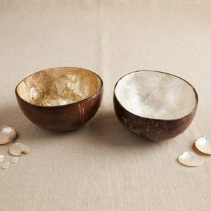 May include: Two round bowls made from coconut shells. The left bowl features a warm, golden-brown iridescent interior lining, while the right bowl has a bright, pearlescent white interior. Both have dark, textured brown exteriors.