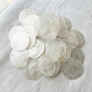 May include: A collection of circular, translucent white shell discs, each featuring two small pre-drilled holes near the edges. These flat, iridescent components exhibit a natural, pearlescent sheen and a smooth, thin texture throughout the pile.