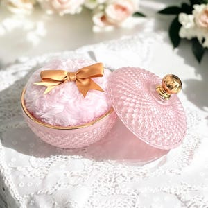 May include: A round, light pink glass powder jar with a textured diamond pattern and a gold-toned rim. The jar contains a soft, fluffy, light pink powder puff topped with a smooth, gold-toned satin ribbon bow. A matching glass lid sits beside it.