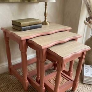 May include: A set of three nesting tables with light wood grain tops and dusty pink painted wooden frames. Each table features four turned legs and a lower shelf with a woven cane insert. The tables are stacked in a descending size order.
