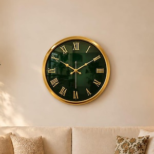 Puede incluir: Un reloj de pared redondo con esfera verde oscuro, números romanos dorados y manecillas doradas a juego, enmarcado en un borde de metal dorado pulido. El reloj está montado en una pared lisa sobre un sofá de color beige.