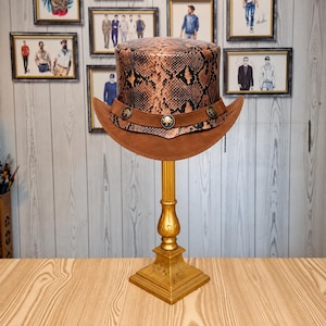 Op de afbeelding: Een hoge hoed met een bruin en zwart slangenleerpatroon op de bol en een brede bruine rand. Een bruine band omcirkelt de basis van de bol, versierd met drie ronde metalen concho's met een reliëf van een bizon.