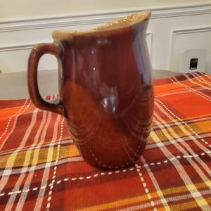 May include: A glossy, dark brown ceramic jug with a smooth, rounded body and a curved handle. The jug features a wide, flared spout at the top rim, with a lighter tan glaze accenting the upper edge and the interior of the pouring area.