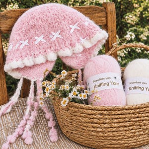 May include: A fuzzy pink knit hat with white ribbon accents and a white pom-pom trim, featuring long braided ties. Beside it, a woven basket contains two balls of Snowflake yarn labeled Knitting Yarn, one pink and one white, with small daisies.