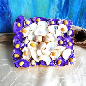Puede incluir: Una bandeja decorativa cuadrada con un mosaico de conchas blancas en el centro, rodeada por un borde de conchas pintadas de color morado vibrante, cada una acentuada con pequeñas piedras lisas de tonos amarillos incrustadas en su superficie.