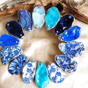 Pode incluir: Um arranjo circular de treze conchas de ostra decorativas, cada uma apresentando padrões únicos em azul e turquesa, cores sólidas ou designs florais. Várias conchas são acentuadas com pequenos strass embutidos e contas de pérolas brancas.
