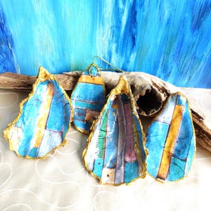 Pode incluir: Quatro conchas de ostra decoradas com um padrão geométrico colorido de riscas azuis, amarelas, verdes e roxas. Cada concha apresenta uma borda pintada em ouro metálico e um acabamento brilhante. As conchas estão dispostas com madeira flutuante.