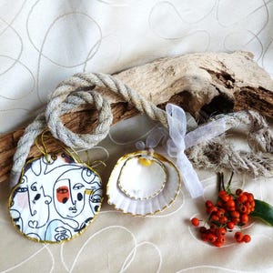 Op de afbeelding: Twee decoratieve schelpen met goudkleurige randen, één met een zwart-witte abstracte gezichtstekening en de andere met een witte binnenkant en een kleine gele kraal, verbonden door een dik beige touw en een wit lint.