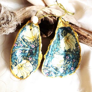 Peut inclure: Deux coquilles d'huîtres décoratives ornées d'un motif floral avec des fleurs jaunes, un feuillage vert et de petites fleurs roses sur fond bleu foncé. Les bords des coquilles sont peints en doré et l'une d'elles possède un cordon doré avec une perle.