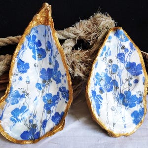 Peut inclure: Deux coquilles d'huître avec un intérieur blanc orné d'un motif floral bleu et des bords peints en doré. Les coquilles sont placées devant un morceau de bois flotté et une corde épaisse de couleur naturelle, formant un objet décoratif.