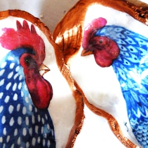 Peut inclure: Deux coquilles d'huîtres peintes avec des coqs détaillés. Chaque coquille présente un intérieur blanc avec une illustration de coq bleu et blanc, dotée d'une crête et d'une caroncule rouge vif. Les bords sont finis en cuivre métallique.