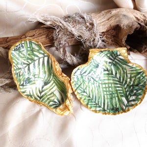 Könnte beinhalten: Zwei dekorative Muscheln mit einem grünen Palmenblattmuster auf der Innenseite und goldfarben bemalten Rändern. Die Muscheln haben die Form einer Auster und einer Jakobsmuschel und zeigen detaillierte botanische Drucke mit grünem Blattwerk.