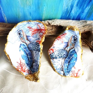 Può includere: Due conchiglie di ostrica con un disegno dipinto di tartaruga marina blu, accenti di corallo rosso e bordi dorati. L'interno di ogni conchiglia mostra un'illustrazione dettagliata di una tartaruga che nuota tra motivi acquatici blu e rossi.