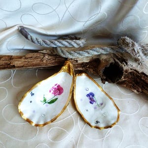 Op de afbeelding: Twee oesterschelpen met goudkleurige randen, elk voorzien van een handgeschilderd bloemmotief. De ene schelp toont een roze bloem, terwijl de andere een paarse bloem heeft. Beide schelpen zijn versierd met kleine, heldere, fonkelende accenten.