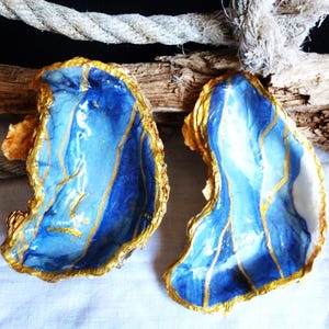 Könnte beinhalten: Zwei Austernschalen, innen mit einem glänzenden blau-weißen Marmormuster bemalt, mit goldfarbenen Rändern und dünnen goldenen Linien, die über die Oberfläche der Schalen verlaufen und ein dekoratives, segmentiertes Aussehen erzeugen.