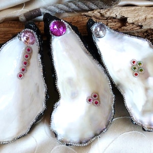 Könnte beinhalten: Drei Austernschalen mit schwarz bemalten Außenrändern und perlmuttfarbenen weißen Innenseiten. Jede Schale ist mit kleinen, funkelnden Strasssteinen in verschiedenen Farben wie Pink, Transparent, Rot und Grün verziert, die in Linien- und Clustermustern angeordnet sind.