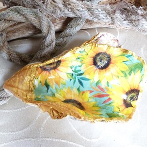 Könnte beinhalten: Eine dekorative Austernschale mit einem Sonnenblumen-Decoupage-Muster, das leuchtend gelbe Blütenblätter und dunkle Mitten auf hellblauem Grund zeigt. Die Ränder der Schale sind mit einem metallisch goldenen Finish bemalt und bilden einen Rand.