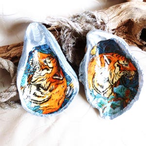 Peut inclure: Deux coquilles d'huîtres peintes avec des illustrations de tigres orange et noirs. L'intérieur de chaque coquille présente une tête de tigre détaillée sur un fond bleu, tandis que les bords extérieurs sont peints en argent métallisé.