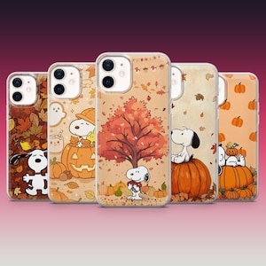 May include: Five clear phone cases featuring Snoopy in various autumn scenes. Designs include Snoopy in a pumpkin, under a fall tree, and surrounded by orange leaves and pumpkins. The cases have a dual-camera cutout and a transparent finish.