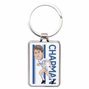 May include: A rectangular silver-toned metal keyring featuring a caricature illustration of a footballer in a white and blue kit. The text "CHAPMAN" is printed vertically in blue capital letters next to the player, set against a white background with blue vertical stripes.