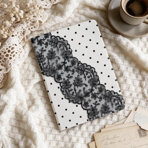 Pode incluir: Uma capa retangular para tablet com fundo branco e um padrão de bolinhas pretas. Uma larga faixa decorativa de renda floral preta é aplicada na diagonal através do centro da capa, exibindo intrincados motivos de folhas e flores.